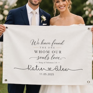 May include: A rectangular white fabric banner with metal grommets in each corner. The text reads: We have found THE ONE WHOM OUR souls love. Song of Solomon 3:4 Katie and Alex with a heart symbol between the names and the date 11.05.2025 below.