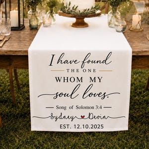 May include: A white fabric table runner featuring black printed text that reads: I have found THE ONE WHOM MY soul loves. Song of Solomon 3:4 Sydney [heart icon] Devin EST. 12.10.2025. The runner is displayed on a wooden table with wedding decor.