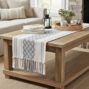 May include: A light wood coffee table with a white and gray patterned table runner. The runner has a diamond design and fringe. A black lantern, books, and a tray with mugs and a bowl are on the table.