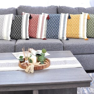 May include: A gray couch with six throw pillows featuring a geometric pattern with tassels. The pillows are in various colors, including black, red, blue, yellow, and green. A wooden tray with a candle, greenery, and wooden beads sits on a gray coffee table with a white runner.