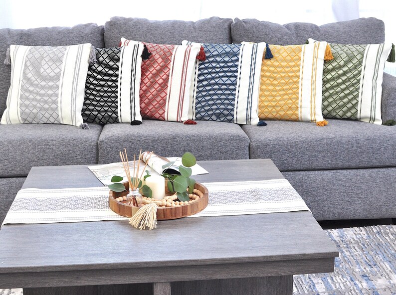 May include: A gray couch with six throw pillows featuring a geometric pattern with tassels. The pillows are in various colors, including black, red, blue, yellow, and green. A wooden tray with a candle, greenery, and wooden beads sits on a gray coffee table with a white runner.