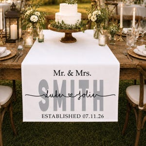 May include: A white fabric table runner featuring black and grey text that reads Mr. & Mrs. SMITH with the names Luke and Jolie written in a cursive script separated by a heart, and the text ESTABLISHED 07.11.26 printed at the bottom.