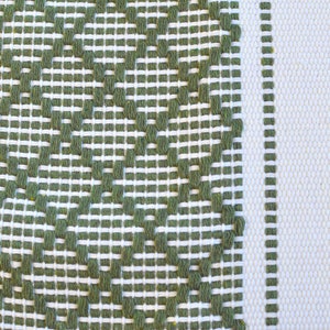 May include: A white fabric with a green woven pattern. The pattern is a series of diamond shapes with a small circle in the center of each diamond.