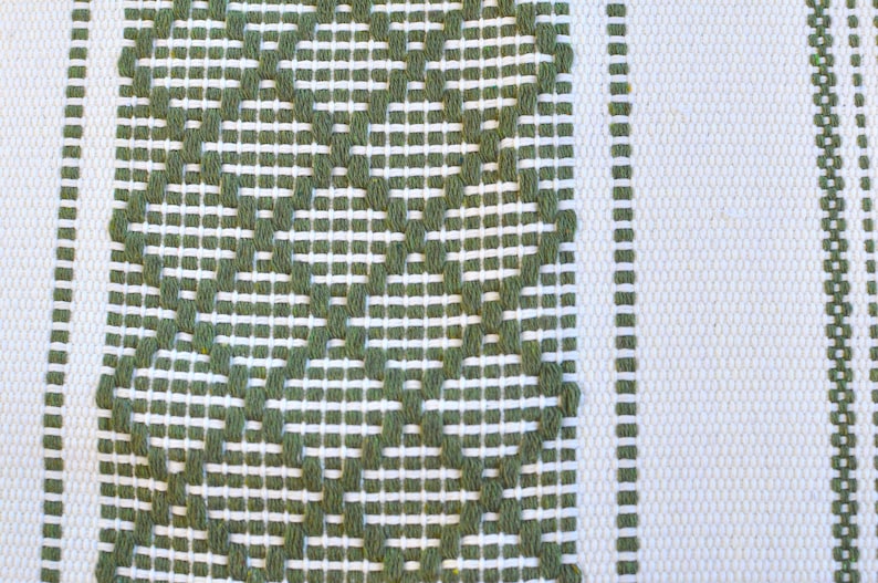 May include: A white fabric with a green woven pattern. The pattern is a series of diamond shapes with a small circle in the center of each diamond.