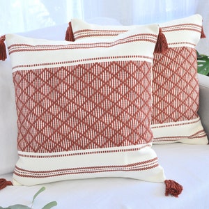 Pode incluir: Duas almofadas decorativas com fundo creme e um padrão geométrico de losangos em vermelho enferrujado. Cada almofada tem uma borla vermelho enferrujado em cada canto. As almofadas estão num sofá branco.
