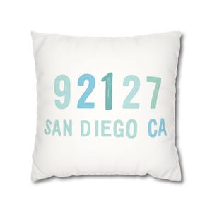 May include: White square cushion featuring the text "92127 SAN DIEGO CA" in shades of blue and green. This cushion is a decorative item, suitable for a home or living area.