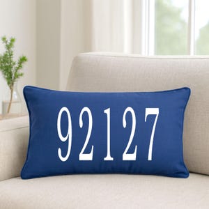 May include: A navy blue rectangular pillow with the white numbers "92127" printed on the front. The pillow is placed on a beige armchair near a window. A small vase with greenery is visible in the background.