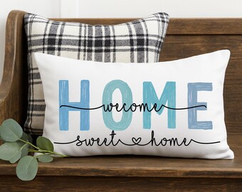 May include: A white rectangular pillow with the word "HOME" in blue, with "welcome sweet home" written below in black script. A plaid pillow is behind it. The pillow is on a wooden surface.