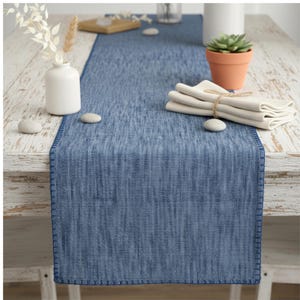 May include: A blue table runner with a textured appearance, featuring a stitched border. The runner is placed on a light-colored wooden table, complemented by a white vase with dried flowers, a small terracotta pot with a succulent, and folded napkins.