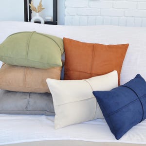 May include: Five decorative throw pillows in various colors and textures. The pillows are arranged in a stack on a white couch. The pillows are made of a soft, suede-like material. The colors of the pillows are green, brown, beige, white, and blue.