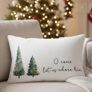 May include: White rectangular pillow with two watercolor Christmas trees and the text "O come let us adore him" in black script. The pillow is on a beige couch with a Christmas tree in the background.
