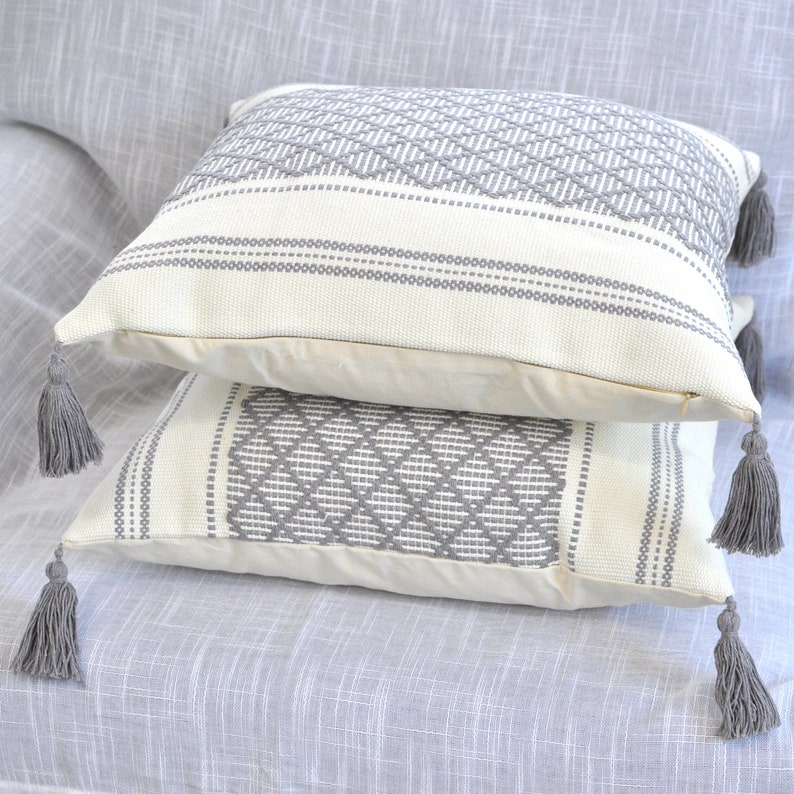 May include: Two decorative throw pillows with a white background and gray geometric patterns. The pillows have tassels on the corners.
