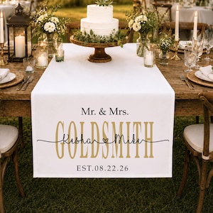 May include: A white fabric table runner featuring black and gold text that reads Mr. & Mrs. GOLDSMITH, Kesha & Mike, and EST.08.22.26. The runner is draped over a wooden table set for a wedding with a tiered cake, floral arrangements, and candles.