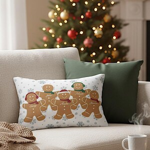 Pode incluir: Uma almofada retangular decorativa com bonecos de gengibre com nomes, flocos de neve e uma árvore de Natal ao fundo. Os bonecos de gengibre têm acessórios coloridos. Duas almofadas verdes também são visíveis.