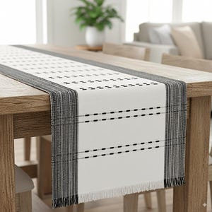 May include: A white table runner with a black and gray geometric design. The runner features a repeating pattern of small black dashes and a fringed edge. The runner is placed on a wooden table.
