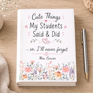 May include: A white hardcover journal featuring the black text "Cute Things My Students Said & Did ...so, I'll never forget" and "Mrs. Carver" with pink heart accents and a colorful floral illustration of pink, orange, and purple flowers at the bottom.