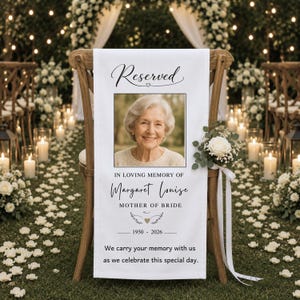 May include: A white memorial sign draped over a wooden chair, featuring a photograph of an elderly woman, the text Reserved, In Loving Memory of Margaret Louise, Mother of Bride, 1950-2026, and a white rose bouquet with a long ribbon.