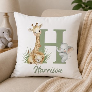 May include: White square pillow with a watercolor illustration of a giraffe, lion, and elephant. A large green letter "H" and the name "Harrison" are also featured. The pillow is on a beige sofa.