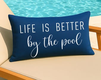May include: A navy blue rectangular pillow with the text "LIFE IS BETTER by the pool" in white. The pillow is on a beige lounge chair next to a pool.