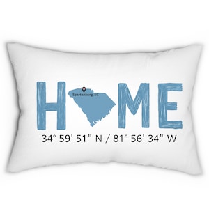 Könnte beinhalten: Weißes rechteckiges Kissen mit dem Wort "HOME" in blauen Blockbuchstaben. Das "O" wird durch eine blaue Karte von South Carolina mit einer Markierung für Spartanburg ersetzt. Darunter zeigt schwarzer Text die Koordinaten 34° 59' 51" N / 81° 56' 34" W.
