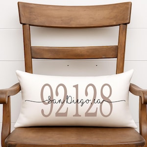 May include: Cream-colored rectangular pillow with the text "92128 San Diego, CA" in taupe and black script. The pillow is placed on a wooden chair with a white background.