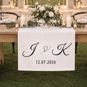 以下が含まれることがあります： 筆記体の黒い文字で「J & K」と印刷され、その下にサンセリフ体のフォントで「12.07.2026」という日付が記された白い布製のテーブルランナー。ランナーはキャンドルや花が飾られた木製のテーブルの上に掛けられています。