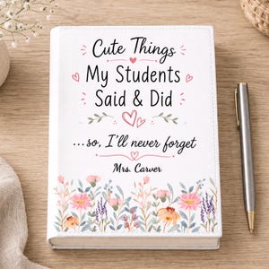 May include: A white journal with the text "Cute Things My Students Said & Did" and "...so, I'll never forget Mrs. Carver." Floral design at the bottom. A silver pen rests on a wooden surface.