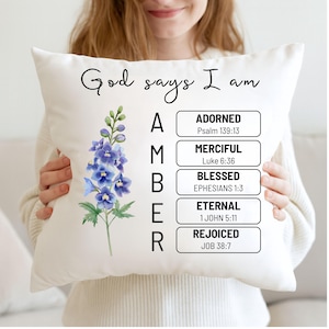 May include: White decorative pillow with the text "God says I am AMBER" in black script. The pillow features a watercolor illustration of blue and purple flowers. The word "AMBER" is followed by a list of positive affirmations.