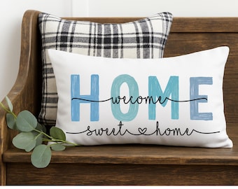 May include: A white rectangular pillow with the word "HOME" in blue and teal, with "welcome sweet home" written below in black script. A plaid pillow is behind it, and greenery is to the left.