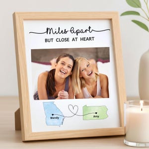 May include: A wooden framed photo print with the text "Miles apart BUT CLOSE AT HEART". The print features a photo of two smiling women, with state outlines and names below. A candle and vase are in the background.