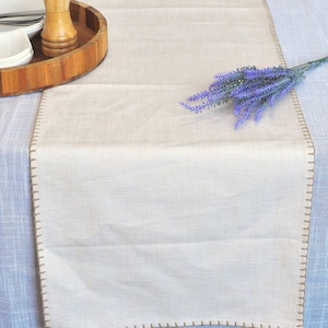 May include: Cream-colored table runner with a stitched border. A wooden tray with a salt shaker and white bowls sits on the left. A sprig of purple lavender rests on the right side of the runner. The runner is on a light blue tablecloth.