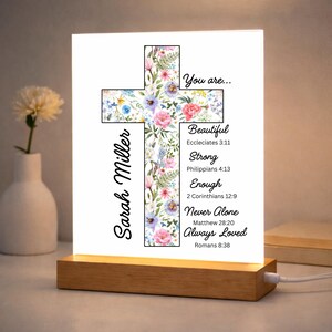 May include: A lighted cross-shaped sign with floral accents and the words "You are... Beautiful, Strong, Enough, Never Alone, Always Loved." The name "Sarah Miller" is printed vertically on the left side. The base is made of wood.