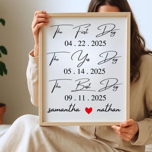 May include: A framed white sign with black calligraphy text. The text reads "The First Day," "The Yes Day," and "The Best Day," each followed by a date. Below, the names "samantha" and "nathan" are separated by a red heart.