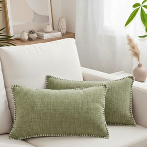 May include: Two rectangular green throw pillows with white stitching on a white sofa. The pillows have a textured fabric and a simple, modern design. The pillows are a muted green color.