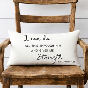 May include: A rectangular white pillow with black text that reads "I can do all this through him who gives me strength Philippians 4:13." The pillow is resting on a wooden chair with a distressed finish.