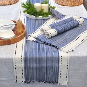 May include: A blue and white striped table runner with matching rolled placemats. The runner has a cream-colored border with a dotted stitch design. The table is set with white plates, silverware, and a wooden tray.