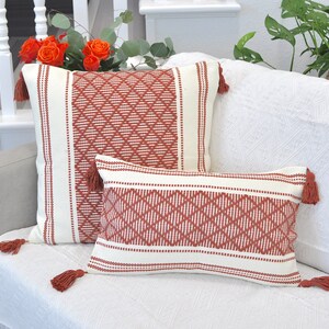 May include: Two decorative throw pillows with a geometric pattern in shades of white and rust. The pillows have tassels on the corners and are arranged on a white couch.