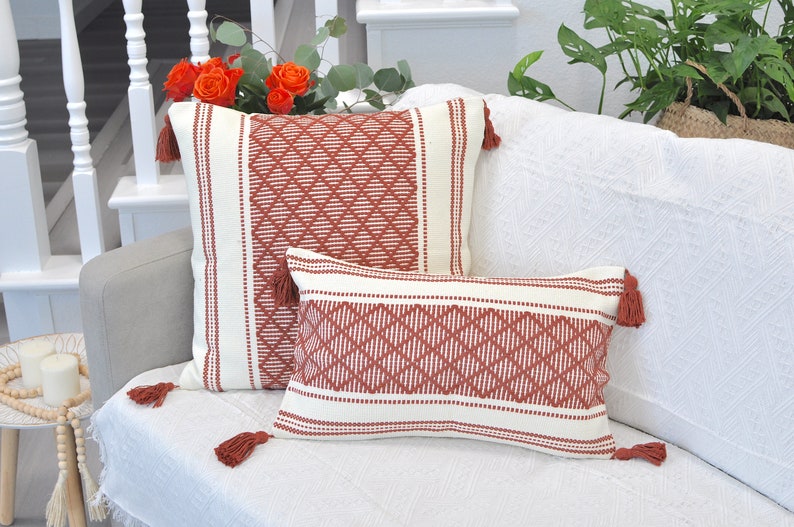 May include: Two decorative throw pillows with a geometric pattern in shades of white and rust. The pillows have tassels on the corners and are arranged on a white couch.