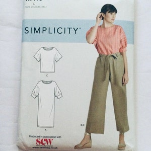 May include: A Simplicity sewing pattern package showing a top and dress design. The package features a photo of a woman wearing a pink striped top and olive green trousers. The text "SIMPLICITY" is clearly visible.