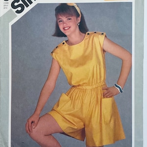 May include: A yellow sleeveless jumpsuit with a round neckline and a drawstring waist. The jumpsuit has two pockets on the front and a button detail on the shoulders.