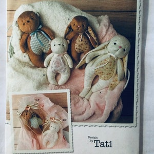 May include: A collection of handmade stuffed animals, including a brown bear with a blue checkered bow, a brown bunny with a floral print, and a white bunny with floral accents. The image also shows a small white bear and a packaged set of the toys.
