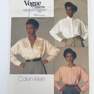 May include: A Vogue Patterns sewing pattern envelope for Calvin Klein designs. The cover features three variations of long-sleeved button-down shirts in white, cream, and peach, paired with skirts. Text includes Vogue Patterns, American Designer, and Calvin Klein.