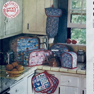 Puede incluir: Una colección de accesorios de cocina, que incluyen fundas para electrodomésticos y agarraderas, con varios patrones. Los artículos se exhiben en una encimera en un entorno de cocina. Se incluye el texto "McCall's Home Center".