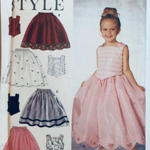 May include: A sewing pattern for a girl's dress featuring a selection of skirt styles. The pattern includes a white skirt with a floral design, a red skirt with a scalloped edge, a grey skirt with a black trim, and a pink skirt with a scalloped edge. The pattern also includes a variety of tops, including a white waistcoat, a black waistcoat, and a floral waistcoat.