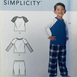 May include: A Simplicity sewing pattern for a boy's pyjama set. The pattern includes a white t-shirt with a blue and white colour block design, a white long-sleeved t-shirt with a grey colour block design, and plaid pyjama trousers.
