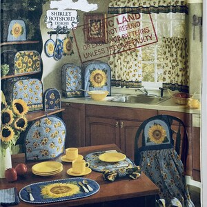 Puede incluir: Escena de cocina con decoración de girasoles. Incluye un calentador de té, fundas para tostadoras, un mantel individual y una funda para silla, todos con diseños de girasoles. Platos, tazas amarillas y un jarrón con girasoles sobre una mesa de madera.