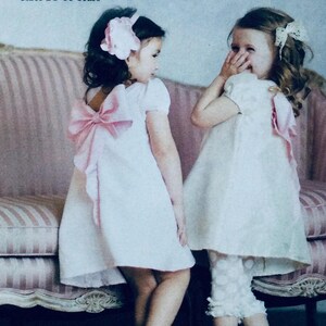 May include: Two young girls in matching white dresses with pink bows and accents. One girl has a large pink bow in her hair, while the other has a white bow. The dresses have short sleeves and a flared skirt. The girls are standing in front of a pink sofa.