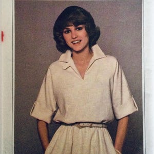 May include: A cream-coloured dress with a collared neckline and short sleeves. The dress features a V-neck, a belted waist, and a skirt that falls to the knees. The sleeves are cuffed, and the dress appears to be made of a lightweight fabric.