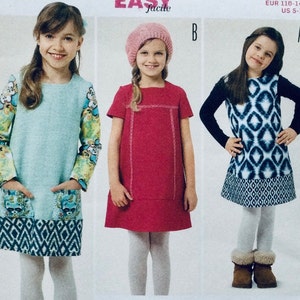 Puede incluir: Tres chicas vistiendo diferentes vestidos. La chica de la izquierda lleva un vestido azul claro con estampado floral en las mangas y estampado geométrico en la parte inferior. La chica del centro lleva un vestido rojo con cuello blanco y un sombrero blanco. La chica de la derecha lleva un vestido blanco y negro con estampado geométrico. Las tres chicas llevan medias blancas.