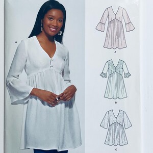 May include: A sewing pattern for a loose-fitting tunic with a V-neckline and 3/4 sleeves. The pattern includes three variations: a white tunic with a button closure, a pink tunic with a lace trim, and a blue tunic with a lace trim.